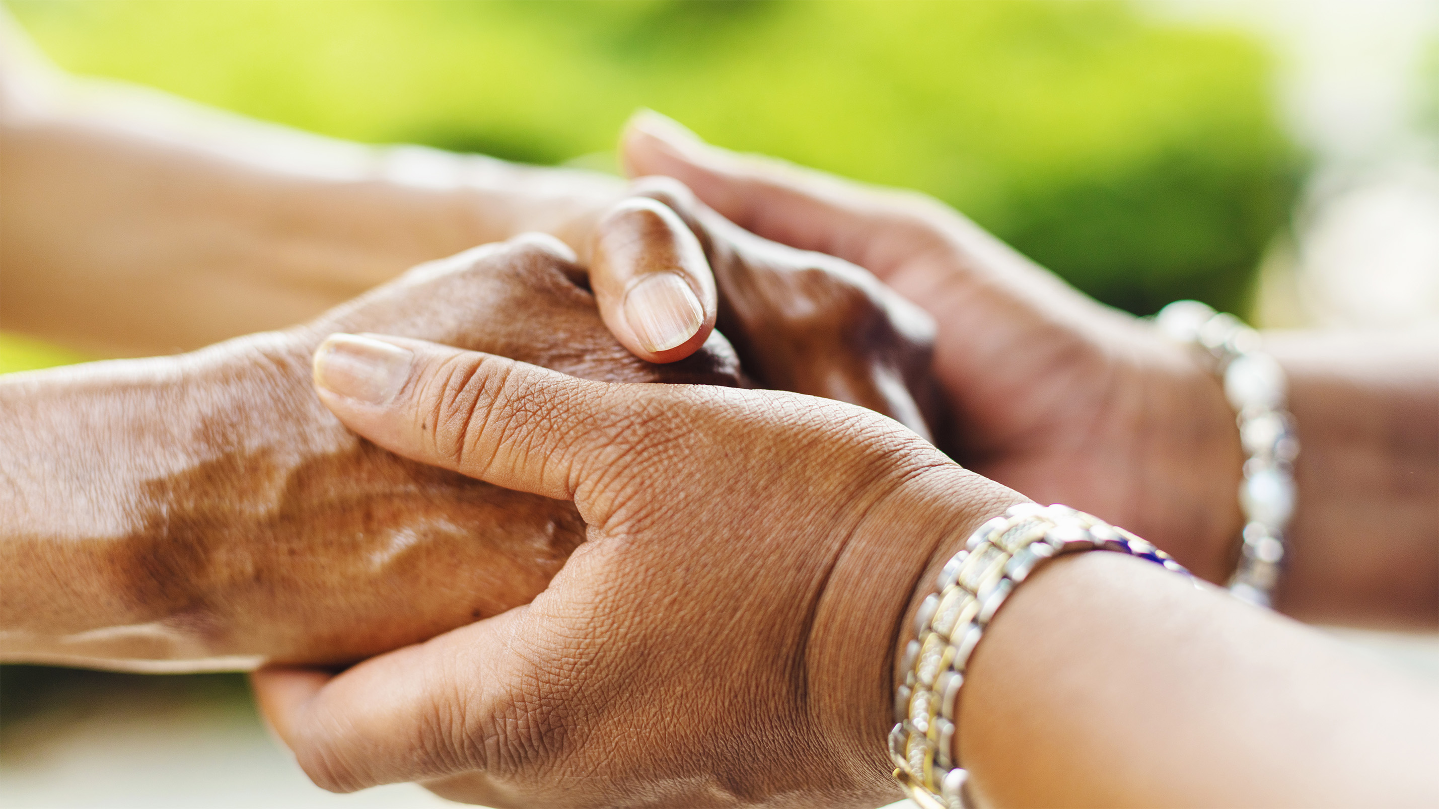 A close-up image of two hands holding each other. One hand is clasping the other gently, with visible wrinkles and a gold bracelet on the wrist. The background is blurred with green hues, suggesting an outdoor setting.